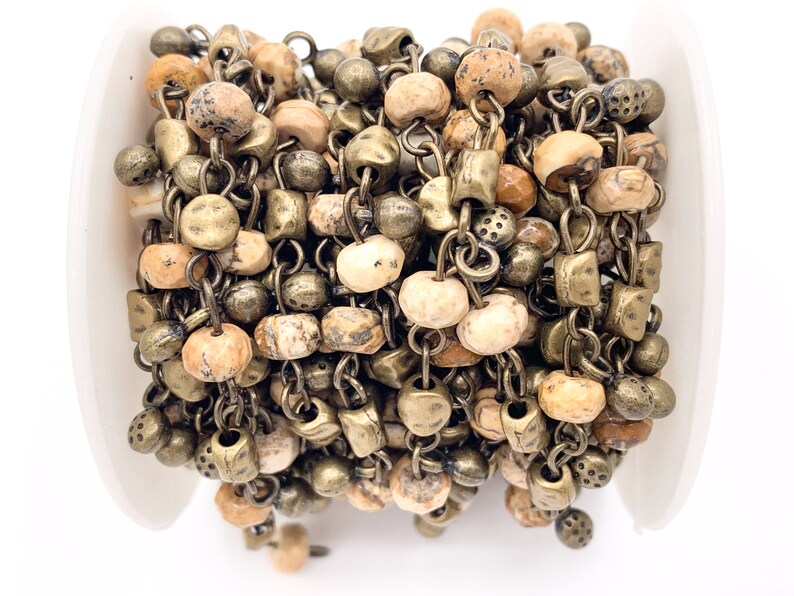 May include: A close-up of a chain made of brown and beige stone beads and antique gold metal links. The chain is wound around a white spool.
