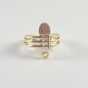 Può includere: Un anello dorato con un design a spilla di sicurezza. L'anello ha tre bande e la spilla di sicurezza è attaccata alla banda superiore.
