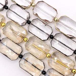 May include: Close-up of a necklace featuring rectangular, faceted beads in clear and amber tones. The beads are outlined in gold and connected by small black and gold accents. The design is geometric and elegant.