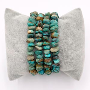 Pu&ograve; includere: Quattro braccialetti in pietra turchese con sfumature di blu, verde e marrone sono presentati su un cuscino grigio. I braccialetti sono realizzati con pietre irregolari e levigate, che mostrano variazioni naturali di colore e consistenza. Un bell'accessorio.