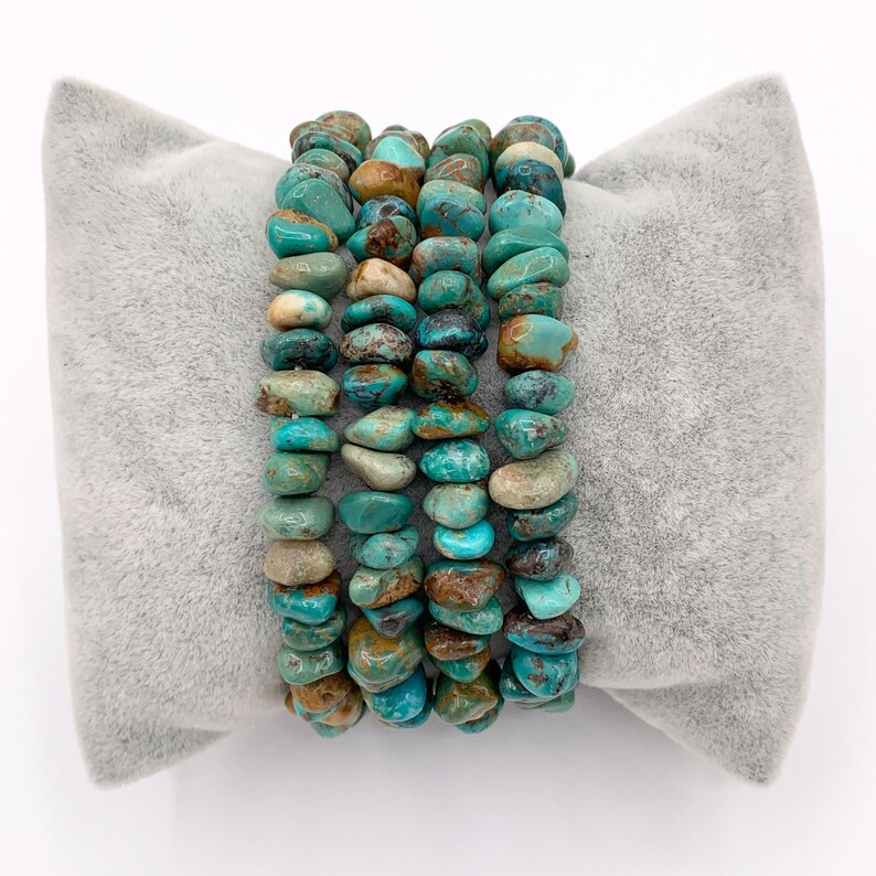 Pu&ograve; includere: Quattro braccialetti in pietra turchese con sfumature di blu, verde e marrone sono presentati su un cuscino grigio. I braccialetti sono realizzati con pietre irregolari e levigate, che mostrano variazioni naturali di colore e consistenza. Un bell'accessorio.