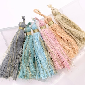 May include: Six colorful tassels with gold thread accents. The tassels are made of soft, fluffy yarn and are available in gray, blue, pink, and beige.