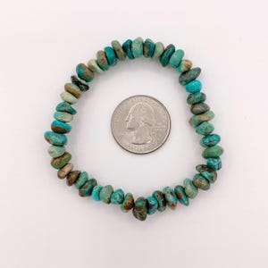 Pu&ograve; includere: Un braccialetto di pietre turchesi con una moneta accanto. Il braccialetto &egrave; composto da piccole pietre turchesi di forma irregolare nelle tonalit&agrave; del blu e del verde. La moneta &egrave; d'argento e ha un profilo di George Washington.