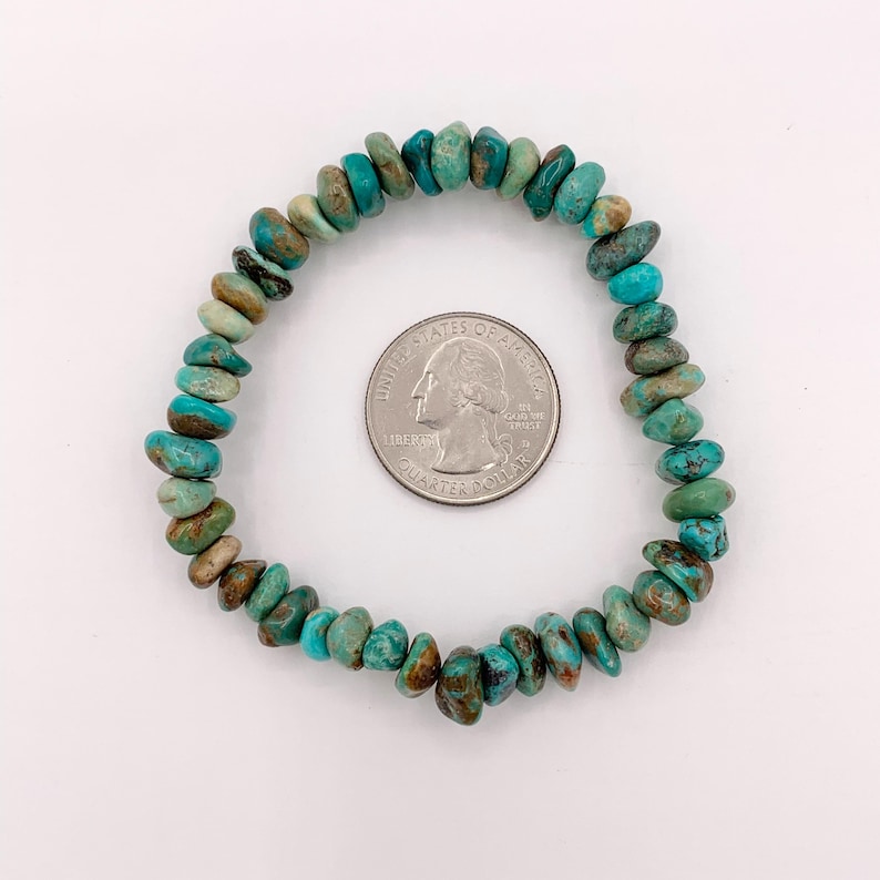Pu&ograve; includere: Un braccialetto di pietre turchesi con una moneta accanto. Il braccialetto &egrave; composto da piccole pietre turchesi di forma irregolare nelle tonalit&agrave; del blu e del verde. La moneta &egrave; d'argento e ha un profilo di George Washington.