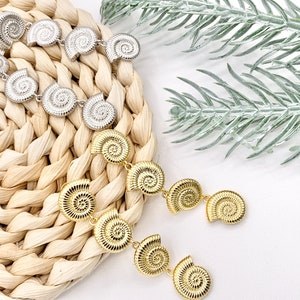 May include: A pair of gold tone earrings with a dangling design featuring multiple spiral shell shapes. The earrings are displayed on a woven straw surface.