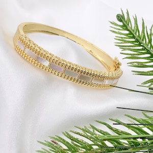 May include: A gold bangle bracelet with white mother-of-pearl accents. The bracelet is made of small beads and has a delicate design.