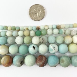 May include: A strand of light green, blue, and white round beads made of amazonite. The beads are approximately 8mm in diameter and are shown next to a US dime for size comparison.