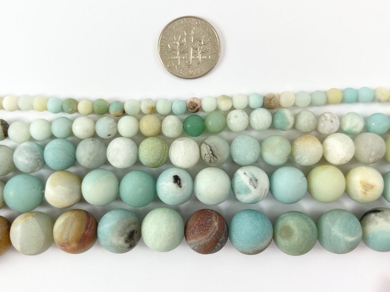 May include: A strand of light green, blue, and white round beads made of amazonite. The beads are approximately 8mm in diameter and are shown next to a US dime for size comparison.