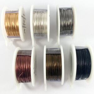 K&ouml;nnte beinhalten: Sechs Spulen Draht in verschiedenen Farben: Gold, Silber, Grau, Kupfer, Bronze und Schwarz. Der Draht ist um eine transparente Kunststoffspule mit einem wei&szlig;en Kunststoffsockel gewickelt.