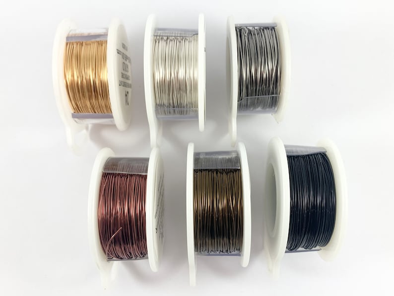 K&ouml;nnte beinhalten: Sechs Spulen Draht in verschiedenen Farben: Gold, Silber, Grau, Kupfer, Bronze und Schwarz. Der Draht ist um eine transparente Kunststoffspule mit einem wei&szlig;en Kunststoffsockel gewickelt.
