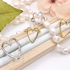 May include: Four gold and silver open heart rings on a wooden display with white pearls.