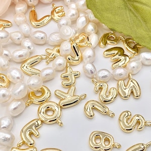 May include: A collection of gold-colored letter and number charms on a strand of white pearls. The charms are in various shapes and sizes, and some have a slightly raised design.
