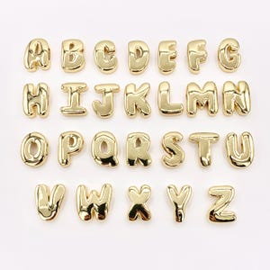 May include: A collection of gold-colored alphabet letters, each with a rounded, bubble-like design. The letters are arranged in rows, showcasing the entire English alphabet from A to Z. The shiny, metallic finish gives them a polished look.