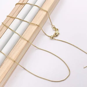 May include: A delicate gold-colored box chain necklace. The chain is displayed on a wooden jewelry display. The necklace features a sliding bead for adjustable length and a lobster clasp closure.
