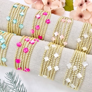 May include: A set of seven gold bracelets with different colored beads and star-shaped charms. The bracelets are arranged in a row on a white fabric background.