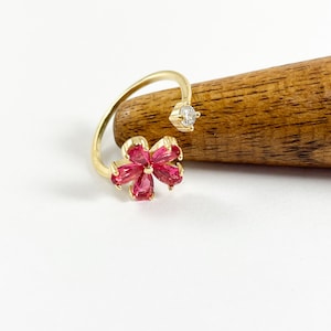 May include: A gold ring with a pink flower design and a single clear stone. The ring is on a wooden surface.