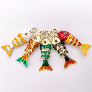 May include: Five colorful fish pendants with gold-tone metal accents. Each fish features a different color scheme, including orange, green, red, black, and yellow, with detailed scale patterns and a small red gem. The pendants are designed for jewelry making.