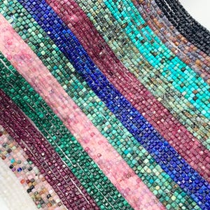 May include: Assortment of square gemstone beads in various colors, including pink, green, blue, and red. The beads are strung on a white background.