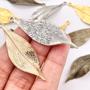 May include: Close-up of several metal leaf charms in various finishes. A gold-toned leaf charm is held in a hand, next to a silver-toned leaf. Other leaves are in gold, silver, and bronze tones. Each leaf has a loop for attaching to jewelry.