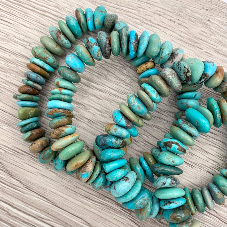 May include: Strands of turquoise gemstone beads in shades of blue, green, and brown. The flat, oval-shaped beads are strung together, forming a necklace or bracelet. The beads have a natural, earthy appearance, with variations in colour and texture.