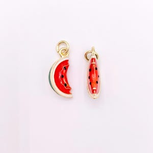 Può includere: Due ciondoli in metallo dorato a forma di spicchi d'anguria. Ogni ciondolo presenta un centro in smalto rosso con piccoli punti neri che rappresentano i semi, un bordo bianco e un contorno verde. Entrambi includono un piccolo anello dorato.