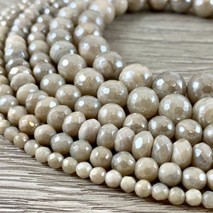 May include: Close-up of several strands of light beige faceted gemstone beads. The beads vary in size, with the largest at the top and the smallest at the bottom. The beads have a subtle sheen and are arranged in a curved pattern.