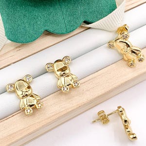May include: Gold-tone bear-shaped earrings with clear crystal accents. The earrings are displayed on a wooden surface.
