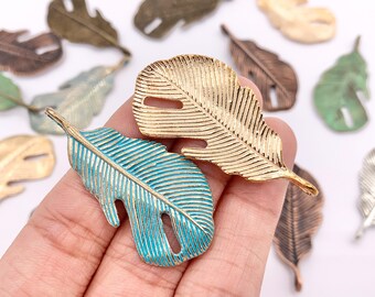 May include: Two metal feather charms, one gold and one teal blue, with a textured design. The charms are on a hand, and there are other metal feather charms in the background.