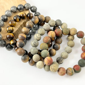 May include: Four beaded bracelets made of different types of stones. The bracelets are in a pile, with the gray and brown bracelets on top. The bracelets are made of tiger's eye, jasper, and other stones.