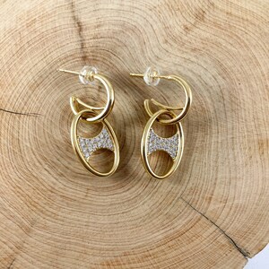May include: A pair of gold hoop earrings with a dangling oval charm. The oval charm is set with small clear stones.
