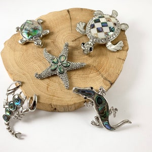 May include: A collection of silver-toned brooches in the shapes of a starfish, turtles, a scorpion, and an alligator, displayed on a wooden slice. The brooches feature abalone shell and rhinestone accents, creating a sparkling, ocean-themed aesthetic.