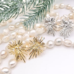 May include: A pair of gold and silver starburst earrings with clear crystals. The earrings are displayed on a strand of white pearls.