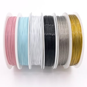 May include: Six circular plastic spools arranged in a row, each wound with a different colored craft wire. The colors from left to right are light pink, light blue, white, black, metallic silver, and metallic gold, all featuring a smooth finish.