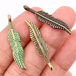 May include: Three metal feather charms in bronze, gold, and green. The charms are detailed with etched lines and a loop at the top for stringing. The feathers are likely for jewelry making or other craft projects.