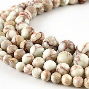 May include: A strand of round, white and brown jasper beads with a marbled pattern. The beads are various sizes and are arranged in a semi-circle.