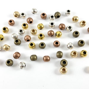 Könnte beinhalten: Eine Sammlung runder Metallperlen in verschiedenen Farben, darunter Silber, Gold und Kupfer. Die Perlen haben eine strukturierte Oberfläche und sind auf einem weißen Hintergrund verstreut.