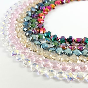 May include: A close-up of a variety of crystal beads in different colors, including clear, pink, champagne, and blue. The beads are shaped like four-leaf clovers and are arranged in a circle.