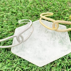 May include: Two gold and silver bracelets with a double-loop design and pave crystals. The bracelets are displayed on a white stone surface.
