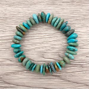May include: A turquoise stone bracelet with a variety of blue and green hues. The stones are irregularly shaped and strung together to form a circular bracelet. The bracelet is displayed on a light wood surface.