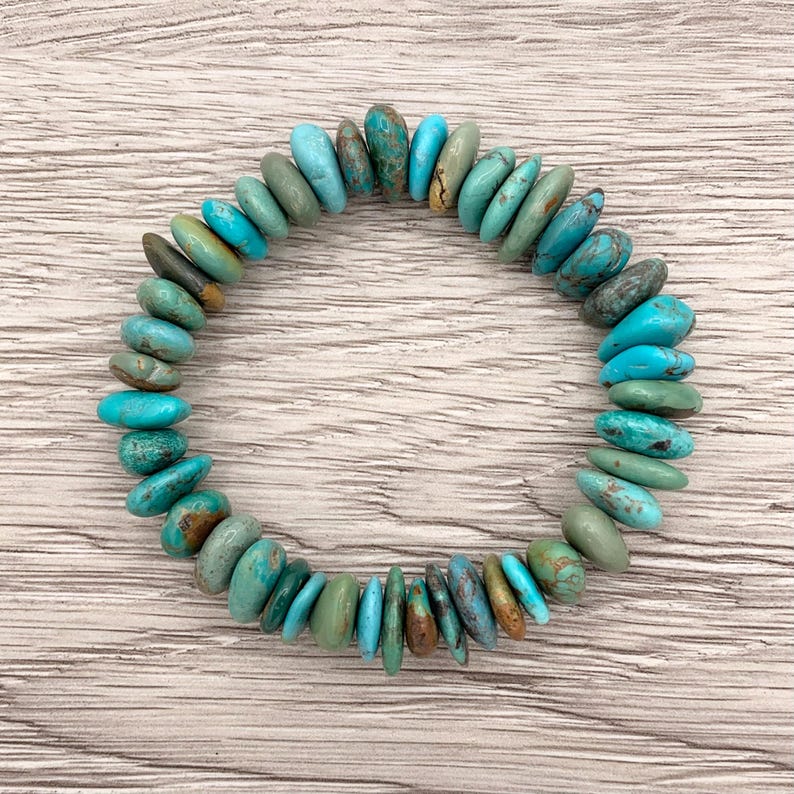 May include: A turquoise stone bracelet with a variety of blue and green hues. The stones are irregularly shaped and strung together to form a circular bracelet. The bracelet is displayed on a light wood surface.