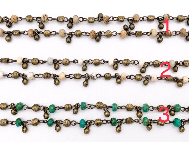 May include: Three strands of chain necklaces with different colored beads. The top strand has brown and white beads, the middle strand has white beads, and the bottom strand has green beads. All strands have small gold-colored charms hanging from them.