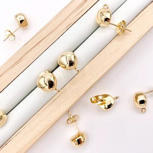 May include: Gold-toned earring findings with a round, slightly domed shape. The findings have a post and a loop for attaching a charm or pendant.