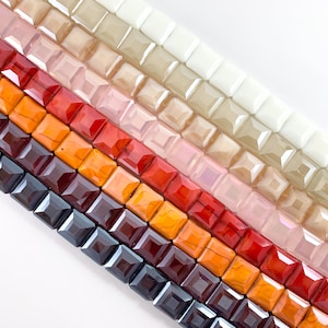 May include: A collection of square glass beads in various shades of red, orange, and brown. The beads are arranged in rows, with the darkest shades at the bottom and the lightest shades at the top.