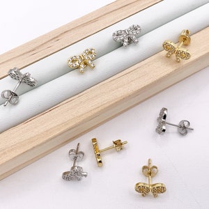May include: A set of six silver and gold bow-shaped earrings with clear cubic zirconia stones. The earrings are displayed on a wooden surface.