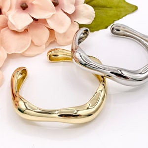 May include: Two minimalist metal bangles, one gold and one silver, with a wavy, organic shape. The bangles are stacked on top of each other.