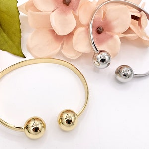 May include: Two open-ended bangles, one gold-toned and one silver-toned, each featuring a spherical accent at the open ends. The gold bangle is on the left, and the silver bangle is on the right. The background includes pink flowers.