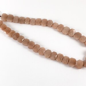 May include: A strand of light brown sunstone beads, each bead is faceted and has a hexagonal shape. The beads are strung on a brown cord.
