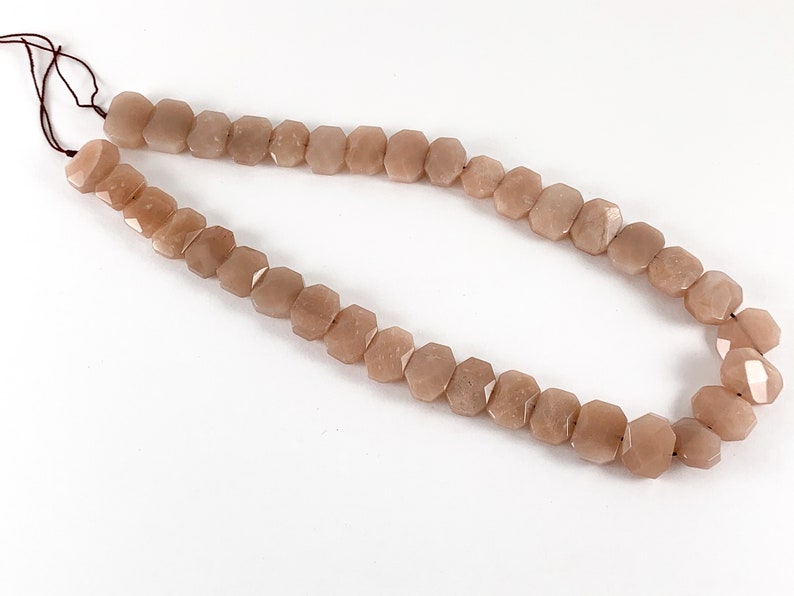 May include: A strand of light brown sunstone beads, each bead is faceted and has a hexagonal shape. The beads are strung on a brown cord.