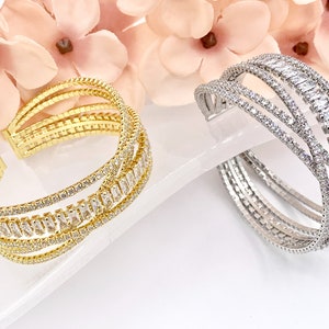 May include: Two gold and silver bangle bracelets with a multi-row design featuring clear cubic zirconia stones. The bracelets are on a white surface.