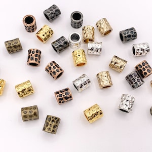 Könnte beinhalten: Sortiment an gehämmerten Metallperlen in verschiedenen Farben, darunter Gold, Silber, Kupfer und Schwarz. Die Perlen sind alle zylindrisch geformt und haben eine strukturierte Oberfläche.